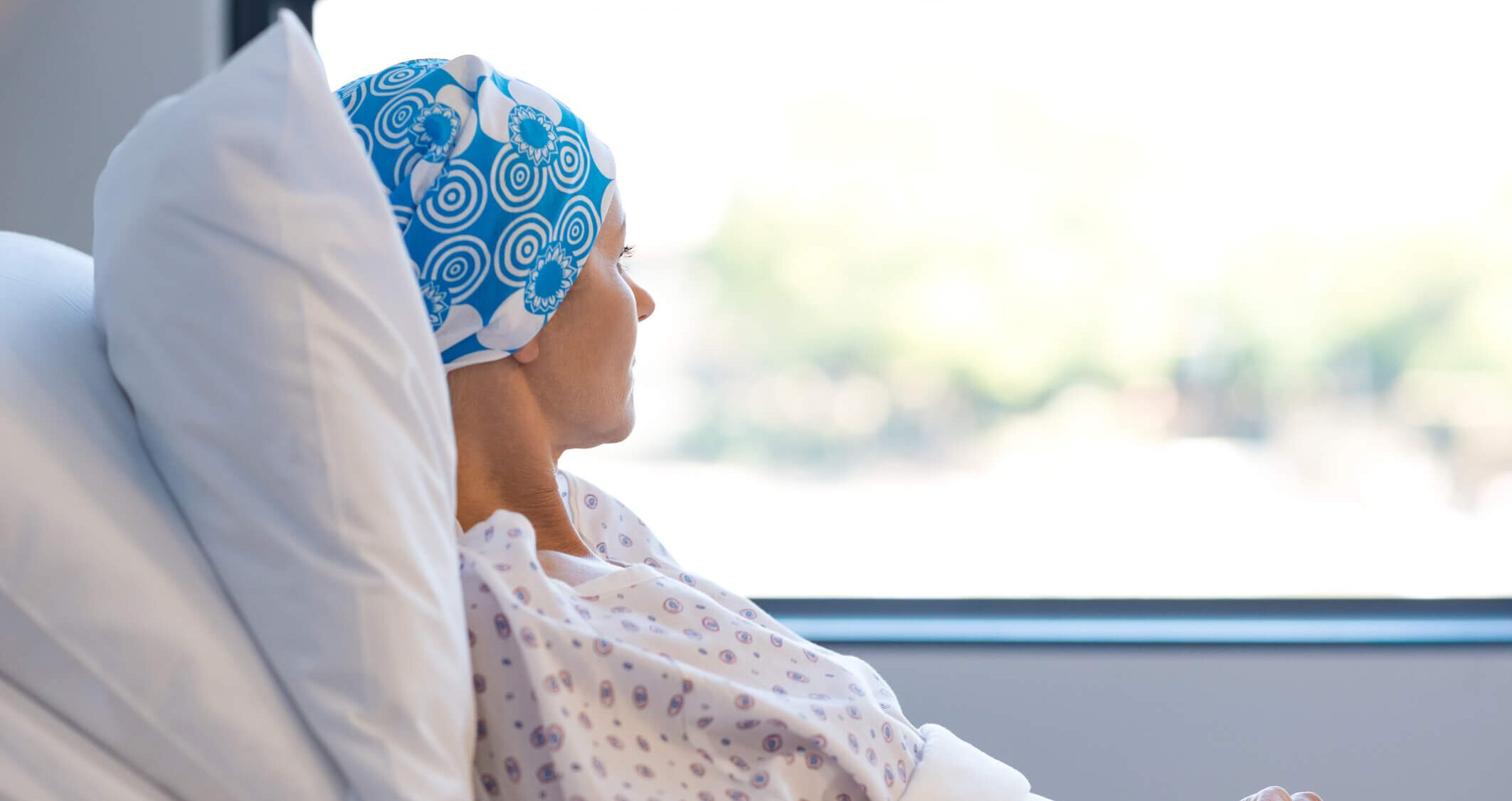 Young woman in bed suffering from cancer. Thoughtful woman battling with tumor looking out of window. Young patient with blue headscarf recovery in hospital on bed.