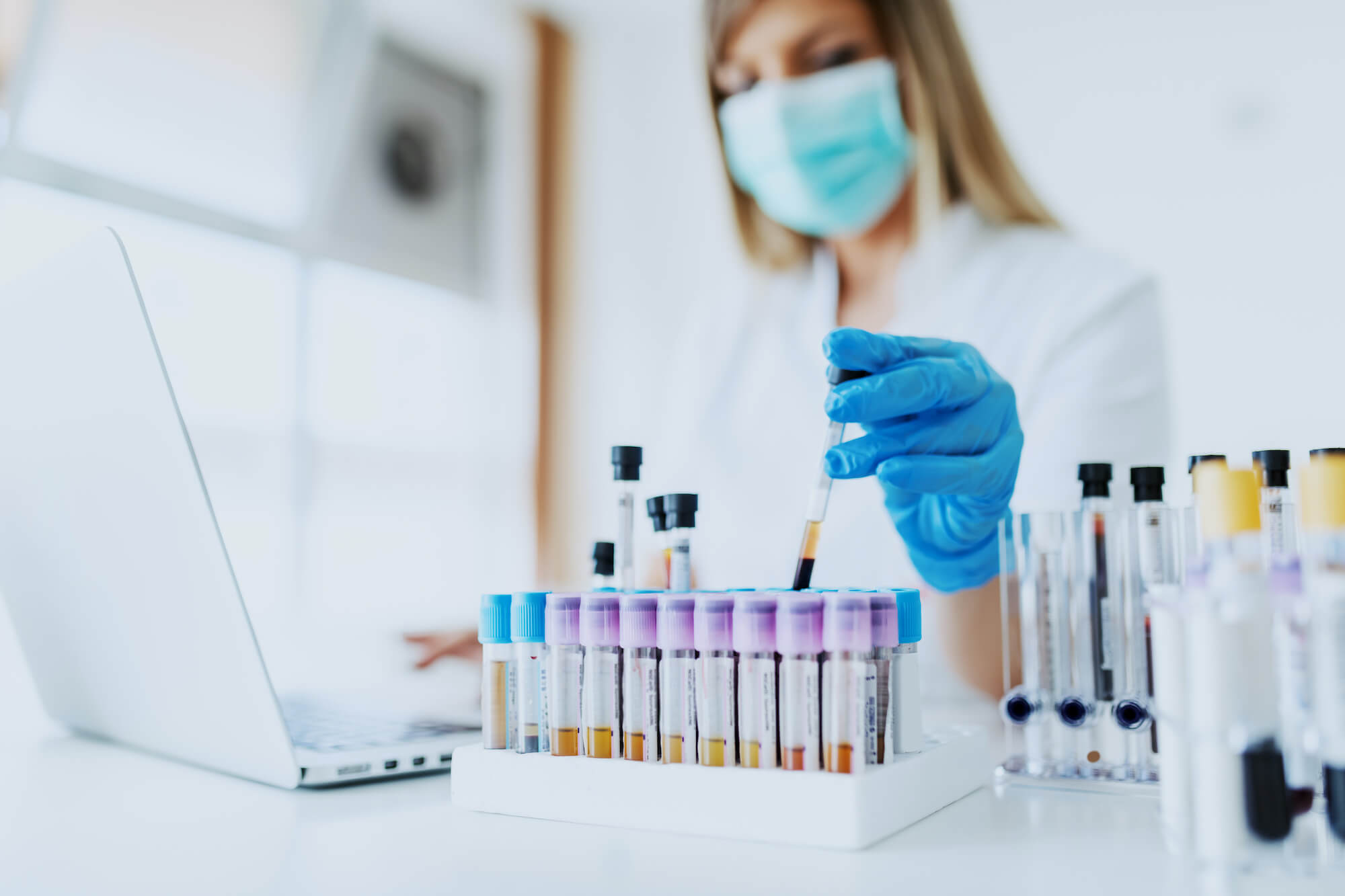 Close up of lab assistant in uniform, with mask and rubber gloves holding test tube with blood sample while sitting on chair and typing on laptop. Selective focus on test tubes. (Close up of lab assistant in uniform, with mask and rubber gloves holdin